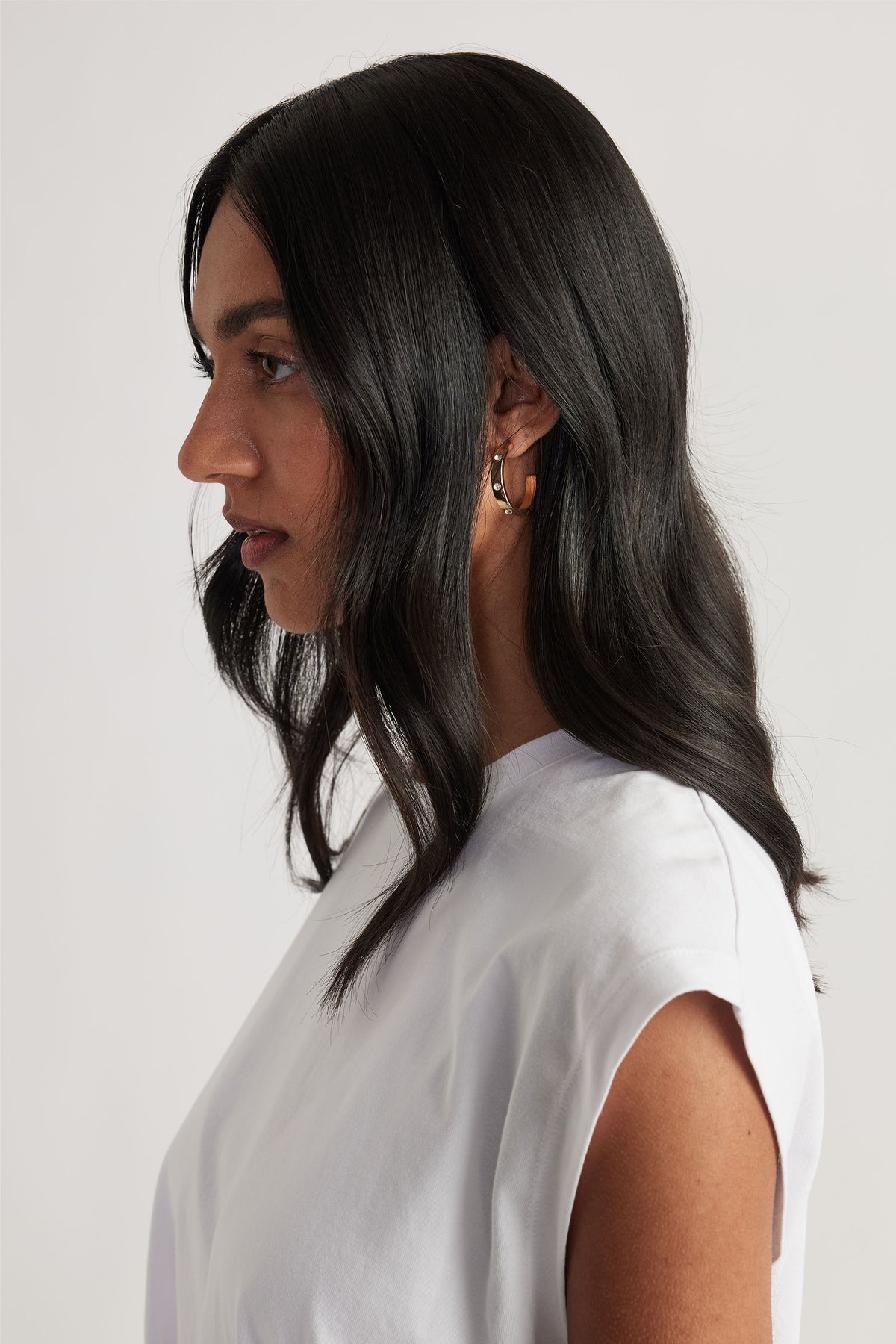 Profile of woman with long dark hair wearing a white sleeveless top and gold hoop earrings with delicate diamanté accents.