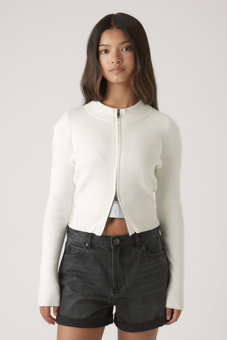 Teen girl wearing a cropped white ribbed zip cardigan paired with rolled black cuff denim shorts for a casual look.