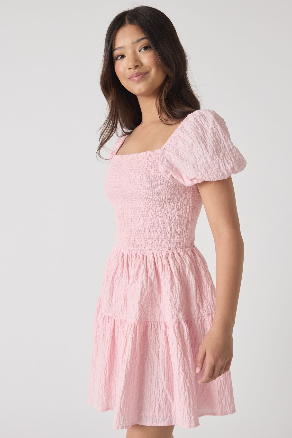 Young woman wearing a textured pale pink mini dress with puff sleeves and tiered skirt, smiling softly at camera.