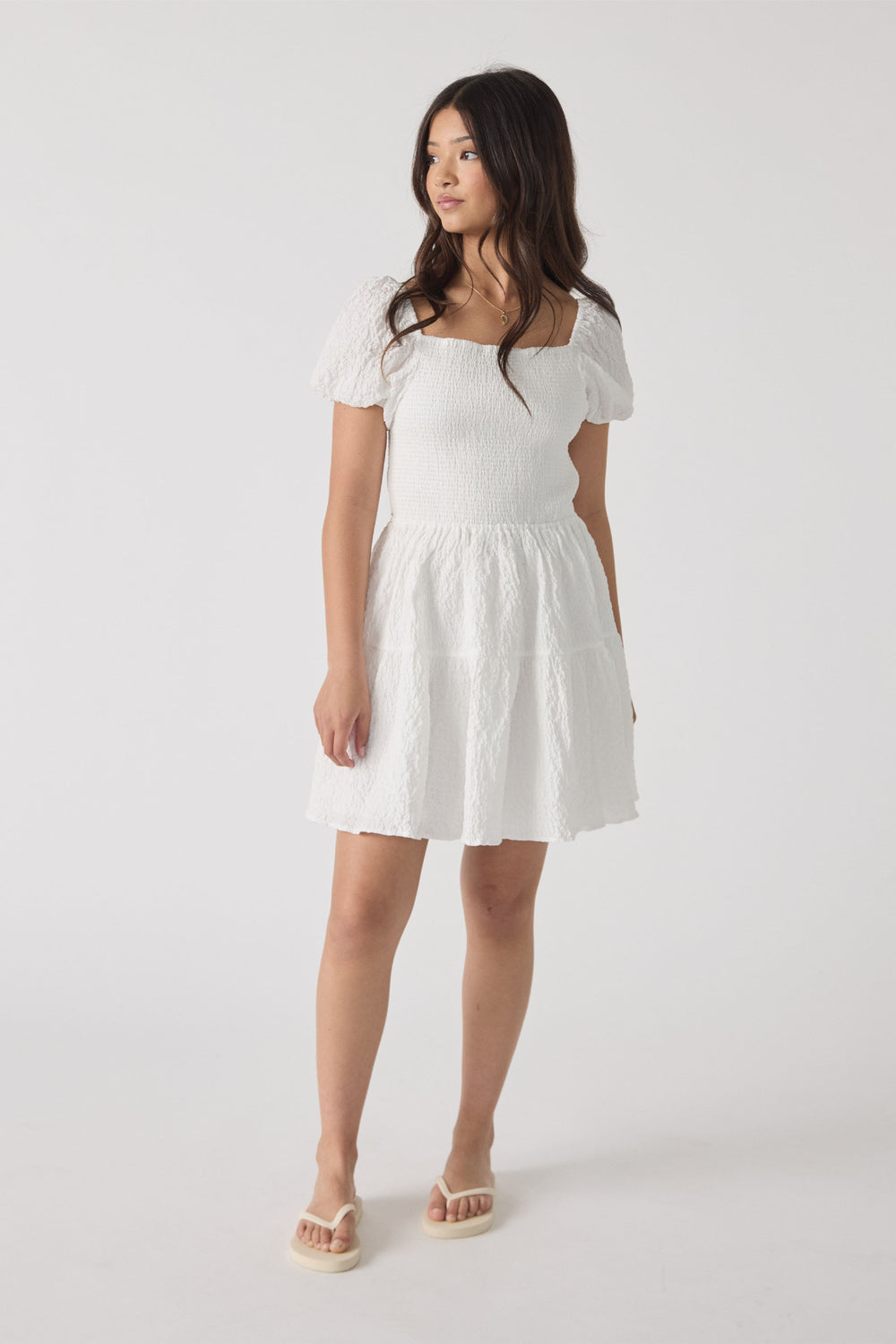 Girl wearing a white textured mini dress featuring puff sleeves and a square neckline, paired with cream sandals.