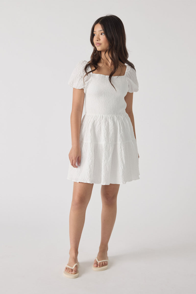 Girl wearing a white textured mini dress featuring puff sleeves and a square neckline, paired with cream sandals.