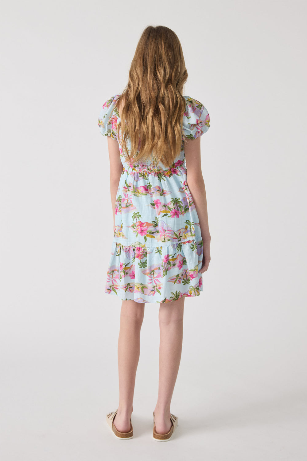Girl wearing a light blue mini dress with pink floral and palm prints, featuring puff sleeves and a tiered hem.