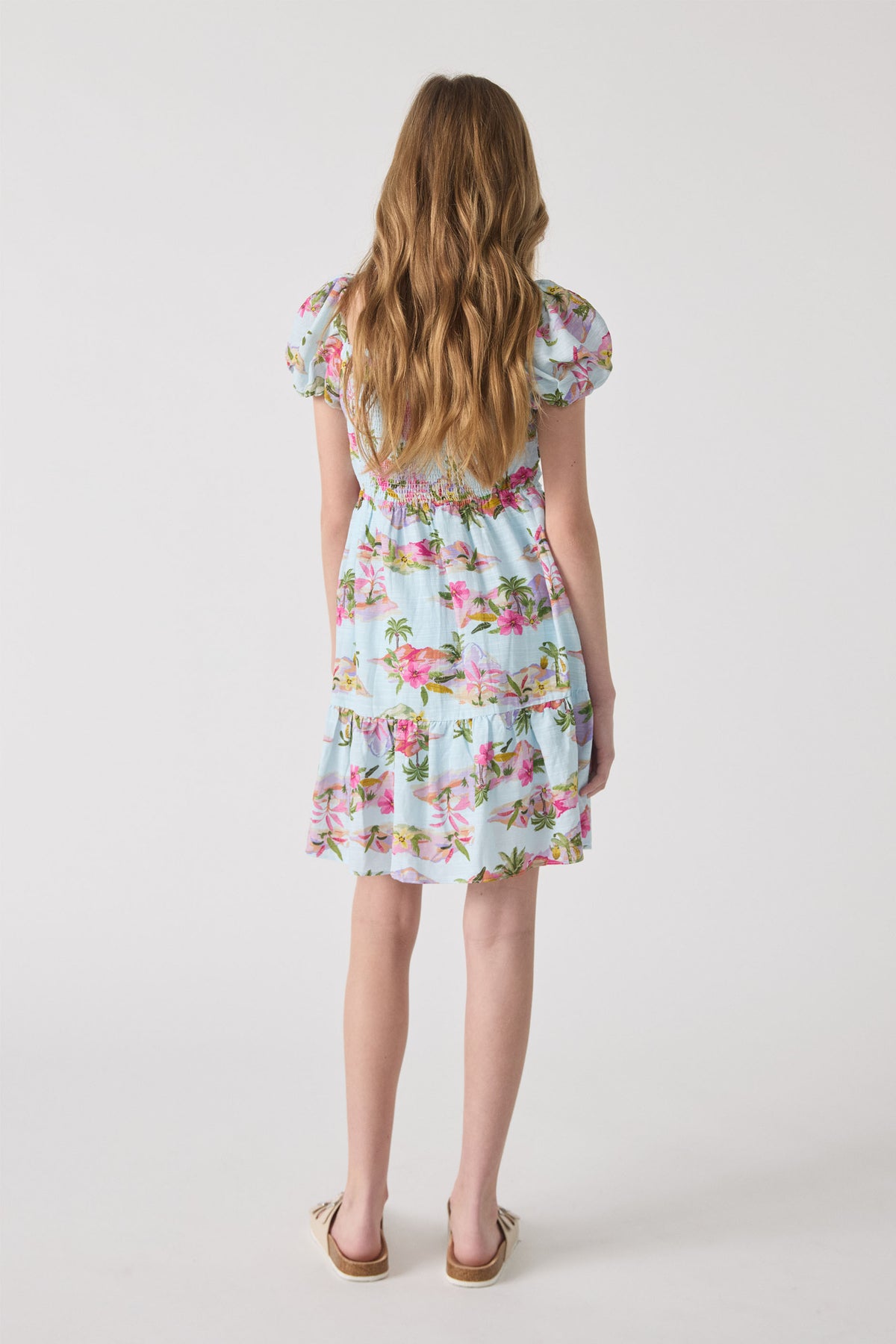 Girl wearing a light blue mini dress with pink floral and palm prints, featuring puff sleeves and a tiered hem.