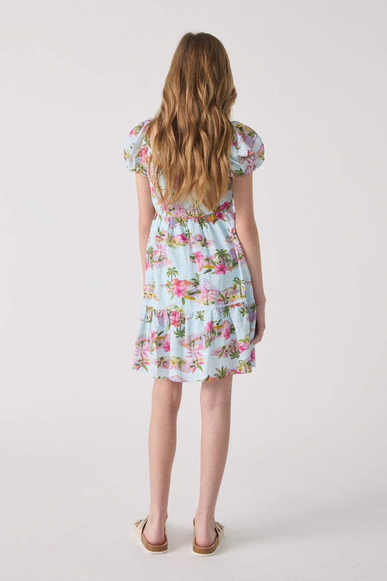 Girl wearing a light blue mini dress with pink floral and palm prints, featuring puff sleeves and a tiered hem.