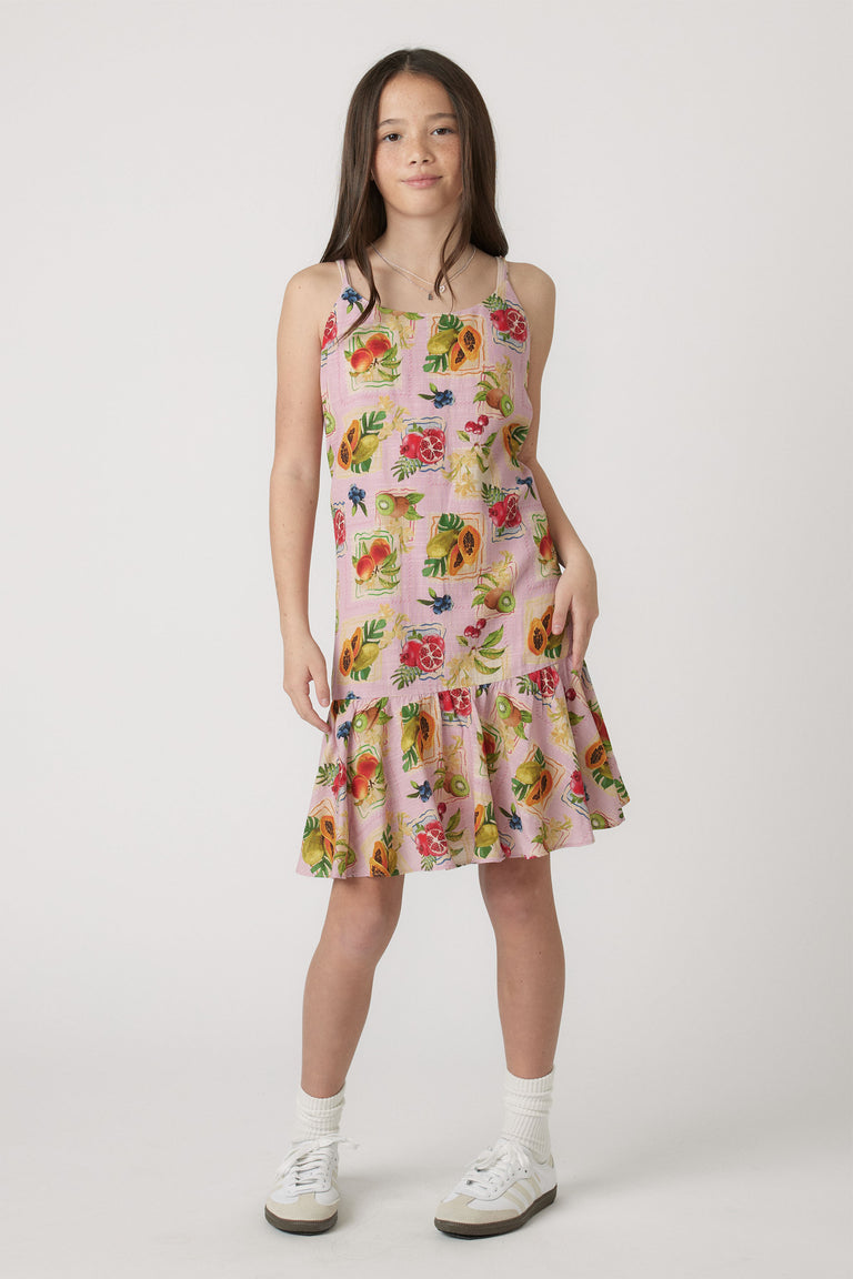 Girl wearing a pink strappy dress with vibrant fruit print and ruffled hem, styled with white socks and sneakers