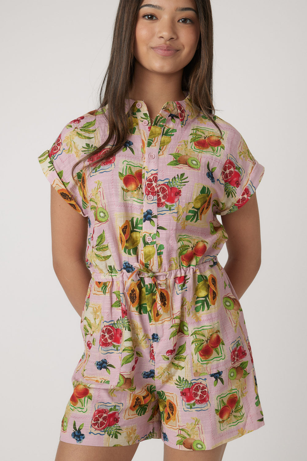 Girl wearing a pink playsuit with a vibrant fruit print, short sleeves, and a belted waist for casual style.