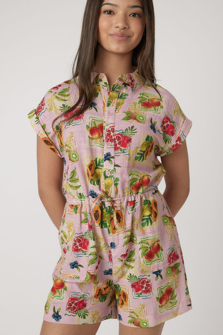 Girl wearing a pink playsuit with a vibrant fruit print, short sleeves, and a belted waist for casual style.