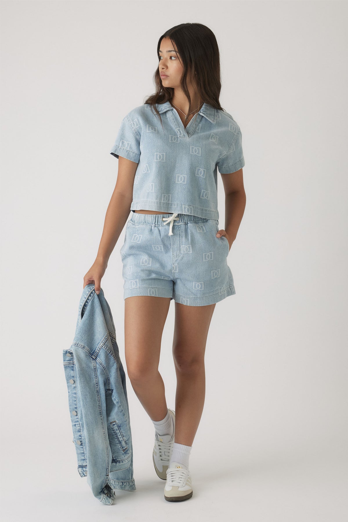 Girl wearing light blue denim shorts and matching short-sleeve top with subtle monogram print, holding a denim jacket.