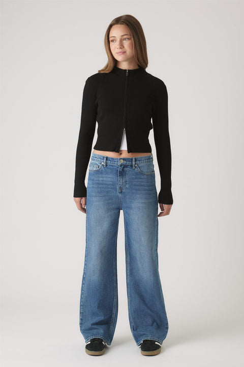 Woman wearing a fitted black zip-up top paired with high-waisted wide-leg blue denim jeans and black sneakers