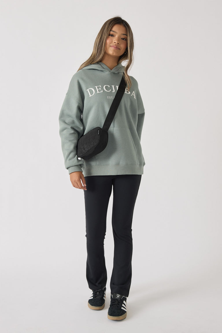 Teen girl wearing a relaxed sage green hoodie with logo print paired with fitted black pants, crossbody bag, and sneakers