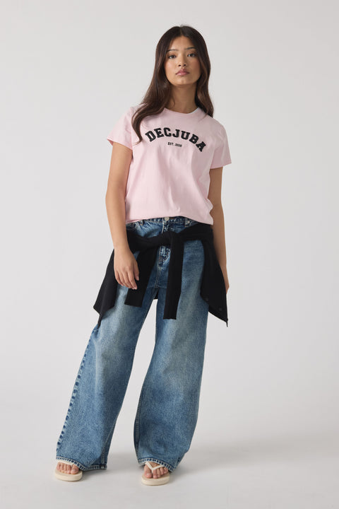 Teen girl wearing a light pink logo tee paired with wide-leg faded blue jeans and a black sweater tied around the waist.