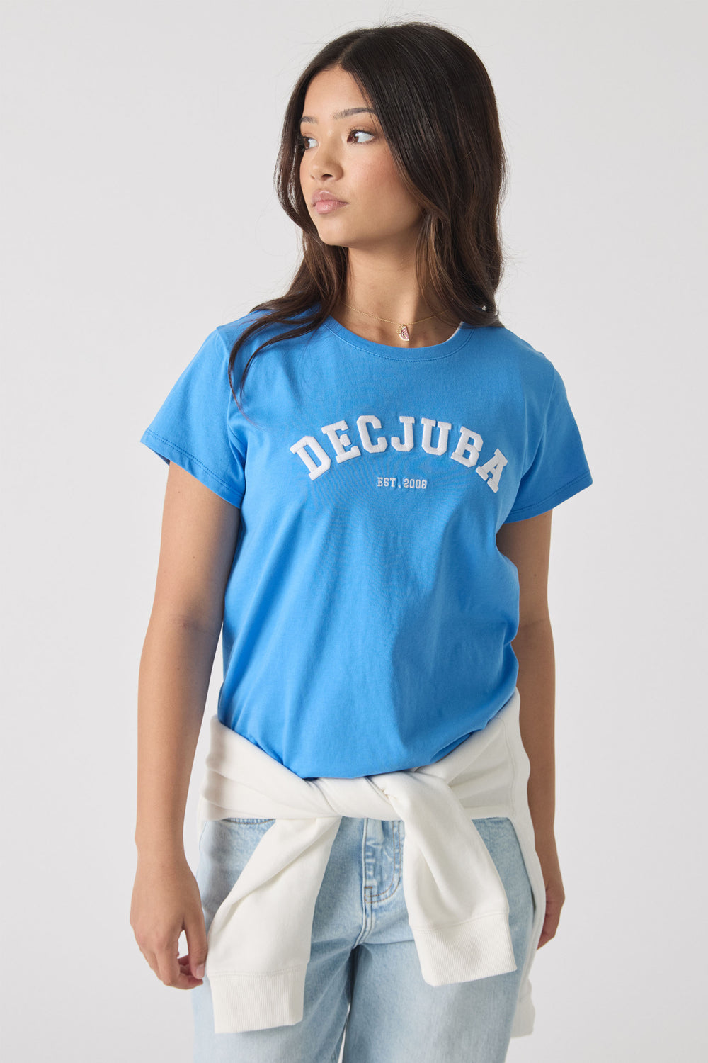 Teen girl wearing a relaxed blue tee with "DECJUBA" lettering, styled with light wash denim jeans and a white sweater tied at waist.