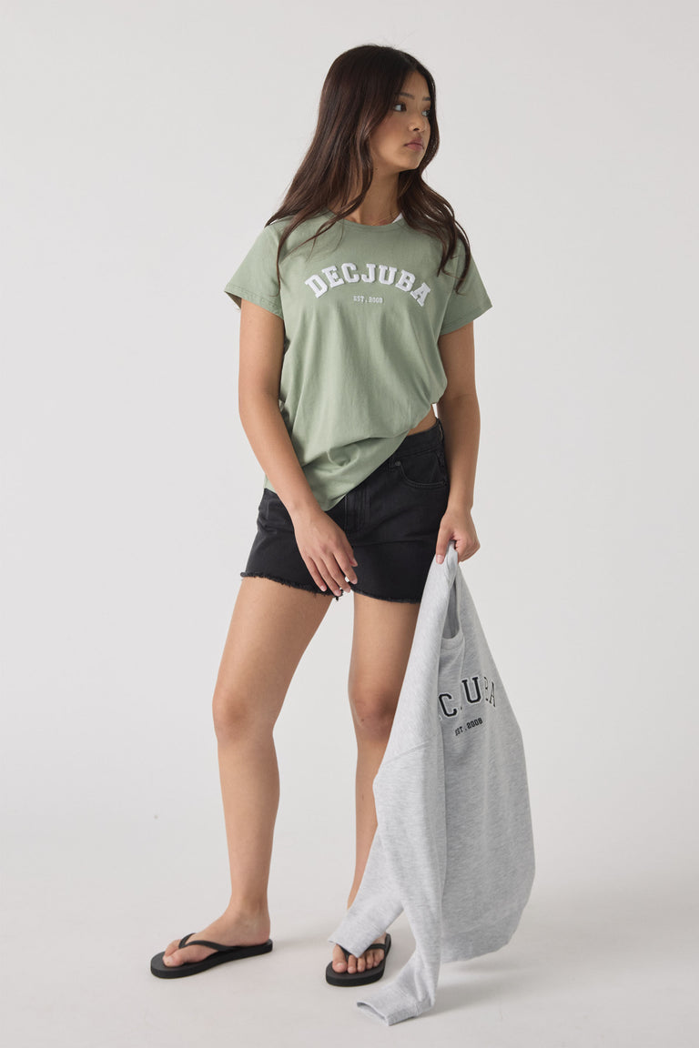 Teen girl wearing a muted green logo tee with black denim shorts and holding a gray sweatshirt by her side.