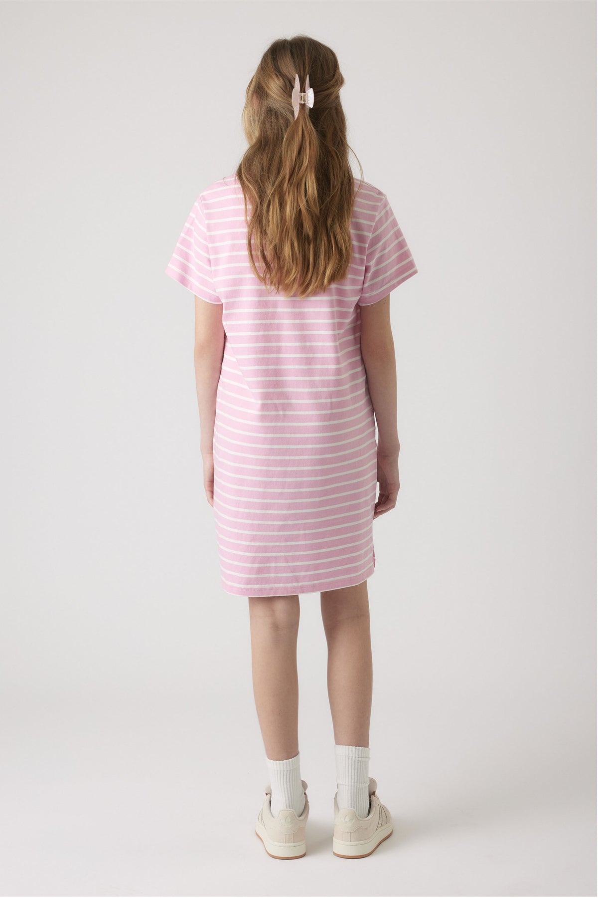 Rear view of girl wearing a soft pink and white striped tee dress paired with white socks and sneakers for casual style