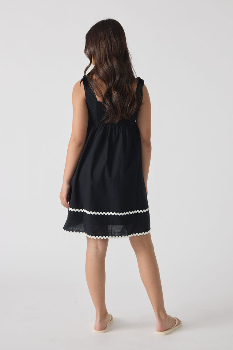 Girl wearing a black sleeveless mini dress with white wavy trim along the hem, shown from behind.