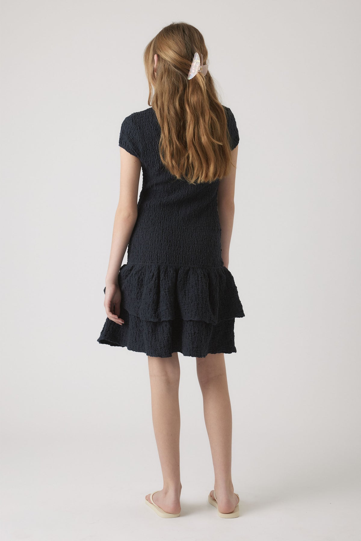 Girl wearing a navy shirred dress with short sleeves and tiered ruffle hem, styled with a pearl hair clip and slip-on shoes