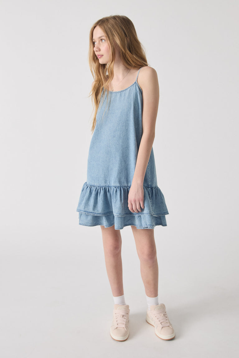Girl wearing a light blue denim dress with thin straps and a double-layered ruffle hem, paired with white socks and sneakers