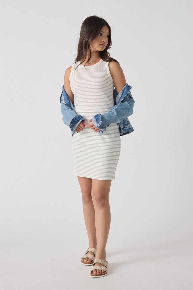 Girl wearing a sleeveless striped tank dress layered with a denim jacket and beige sandals, standing against a plain background.