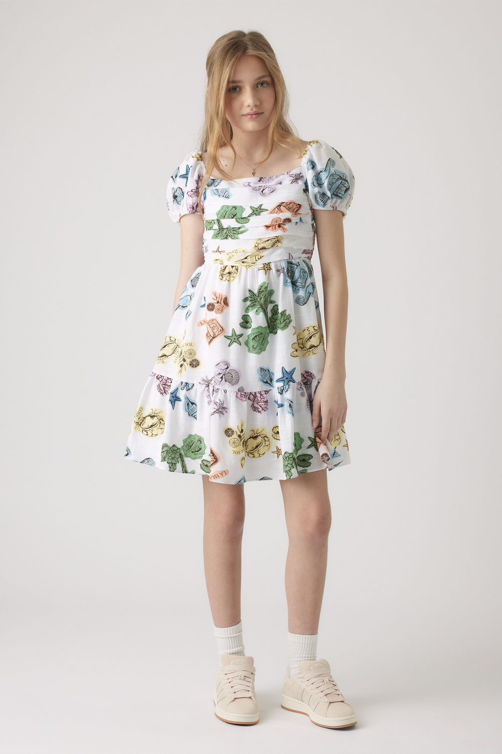 Girl wearing a white mini dress with colorful shell and starfish prints, puff sleeves, and a tiered skirt, paired with sneakers.