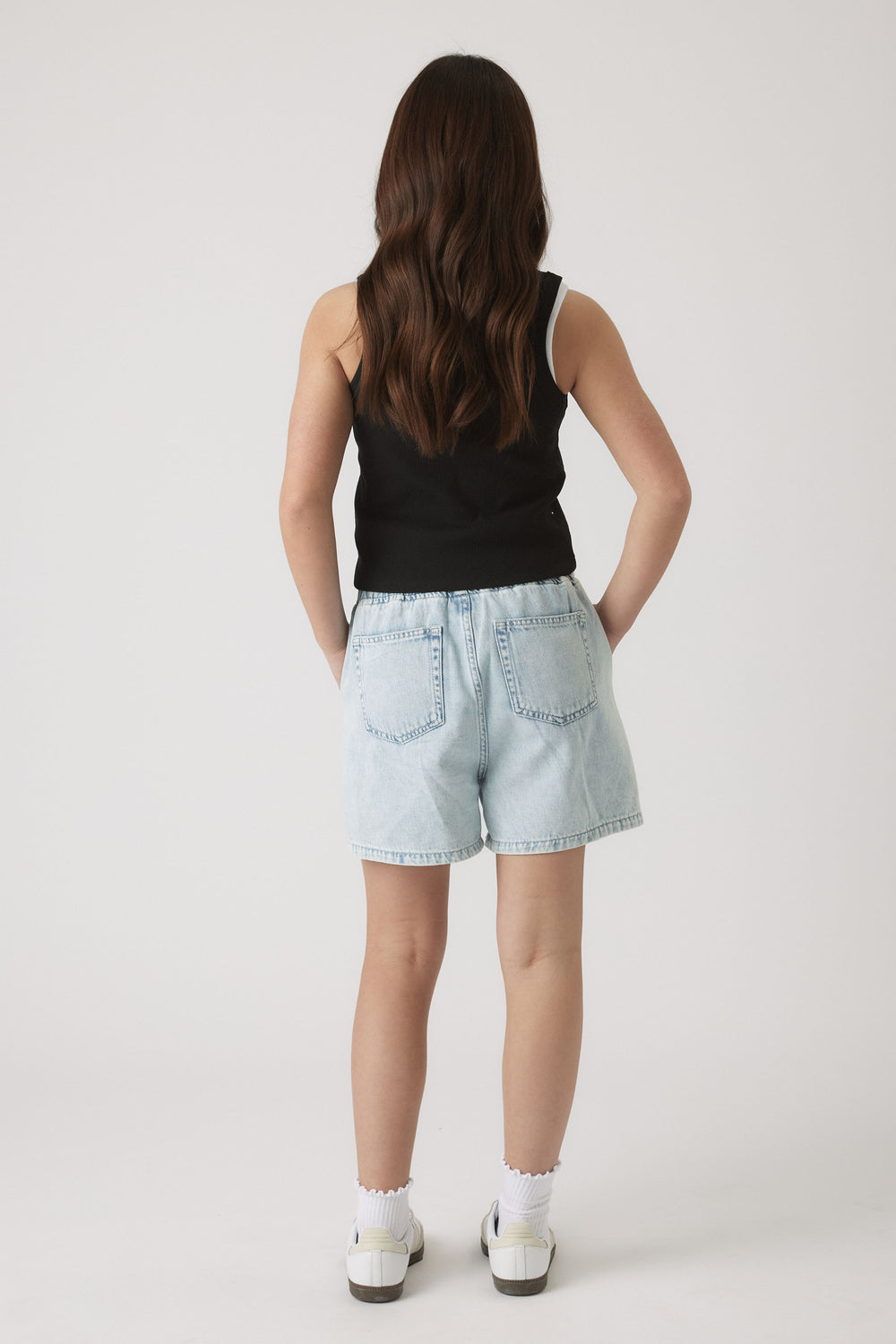 Girl seen from behind wearing a black tank top paired with light wash casual denim shorts and white socks with sneakers.
