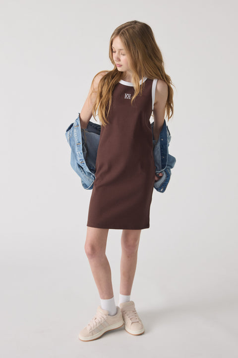 Girl wearing a sleeveless chocolate brown monogram dress layered over a white top, paired with white sneakers and a denim jacket.