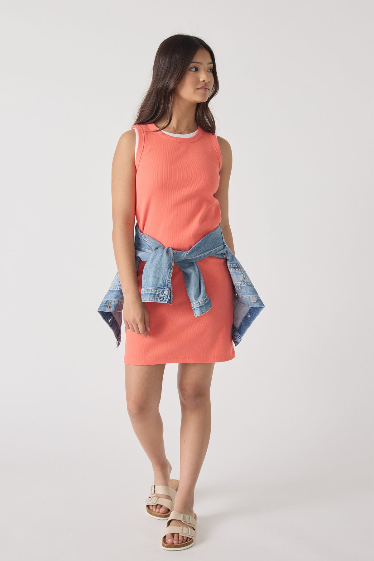 Girl wearing a coral ribbed tank dress with a denim jacket tied around the waist and beige sandals walking forward