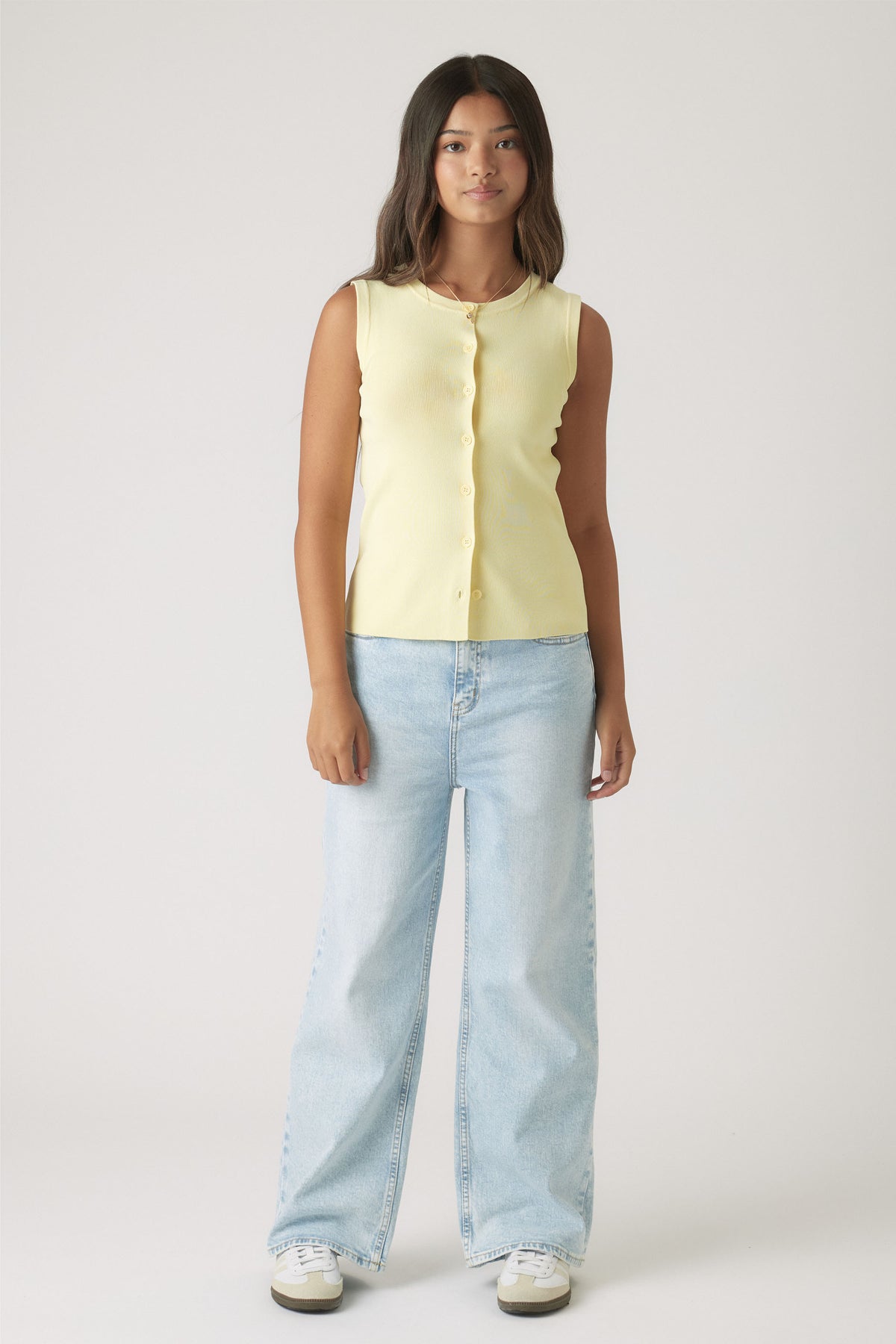 Girl wearing a soft yellow sleeveless knit vest with buttons paired with light wash wide-leg denim jeans and sneakers