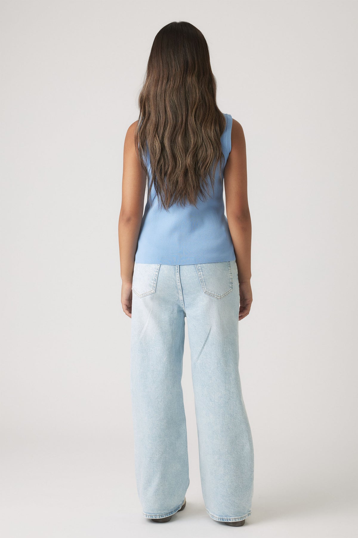 Girl with long wavy hair wearing a dusty blue sleeveless knit vest paired with light wash wide-leg denim jeans