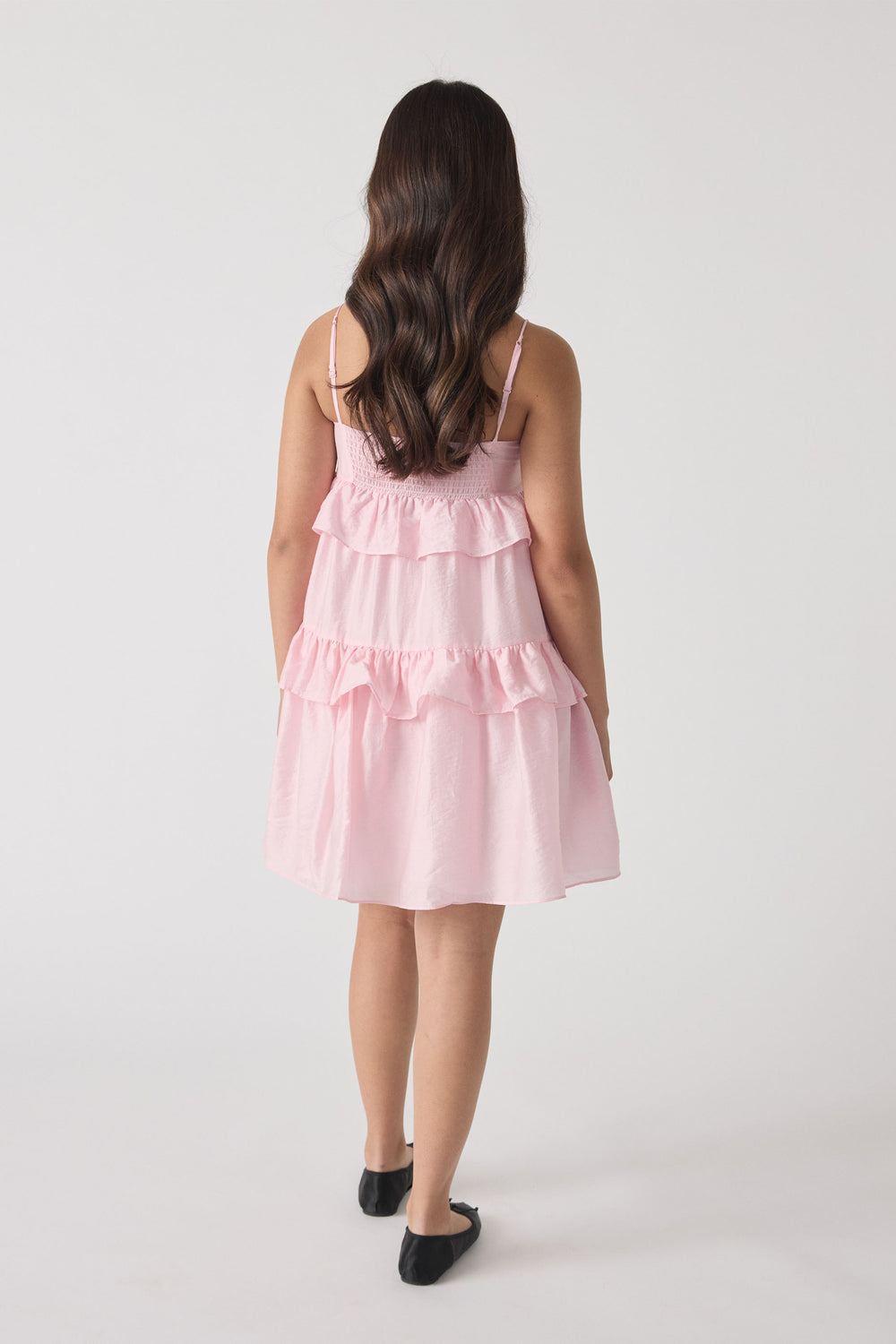 Young girl in a soft pink tiered ruffle dress with thin straps and black ballet flats, shown from behind.