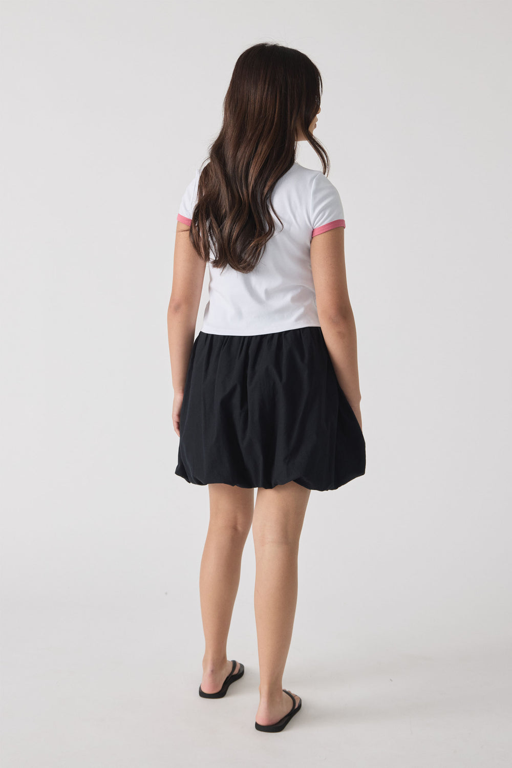 Girl wearing a white short-sleeve top with pink trim paired with a black bubble mini skirt and black slip-on sandals, shown from behind.