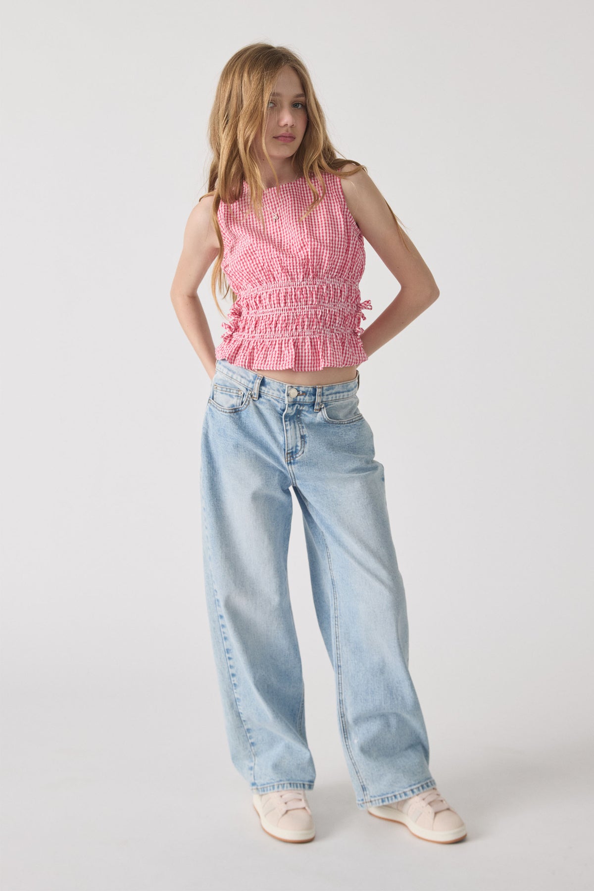 Girl wearing a sleeveless pink gingham tie top with smocked waist, paired with light blue wide-leg jeans and sneakers