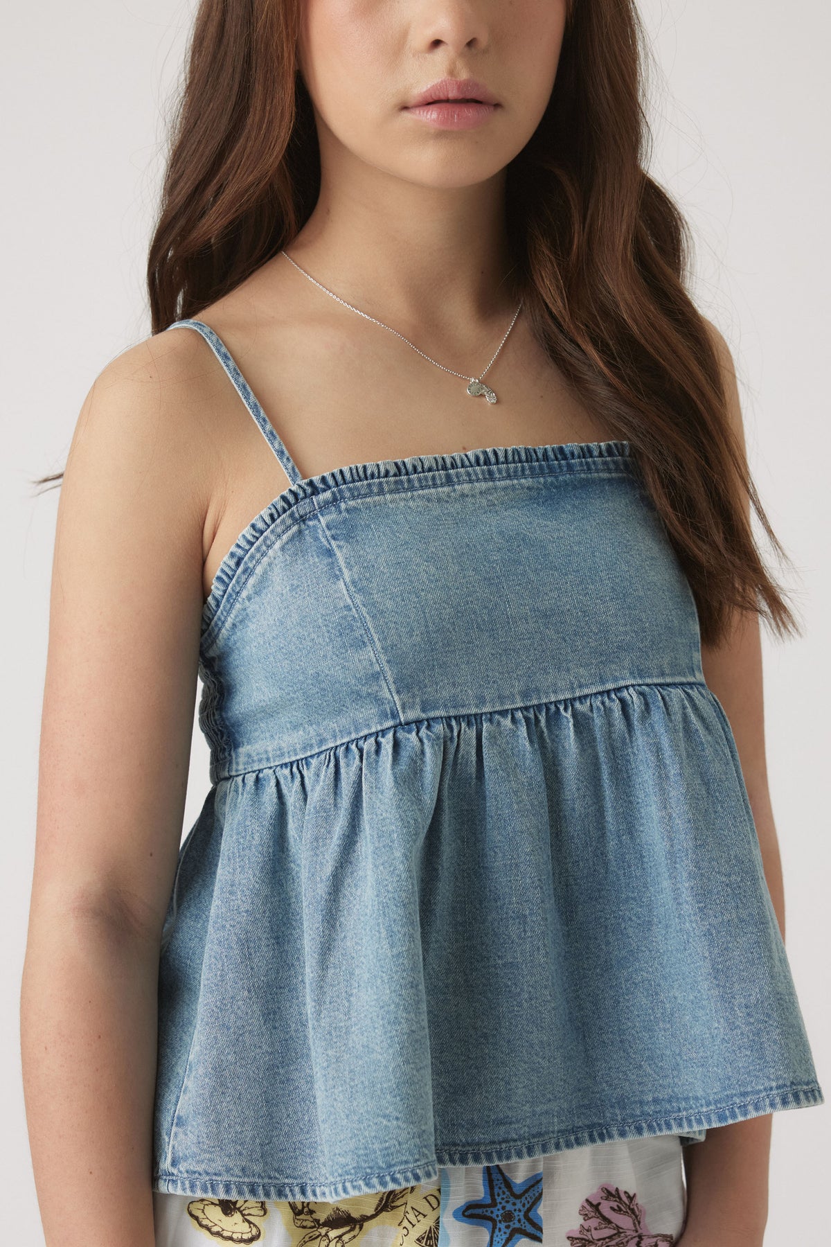 Girl wearing a midwash denim top with thin straps and a gathered, loose fit hem for a casual look