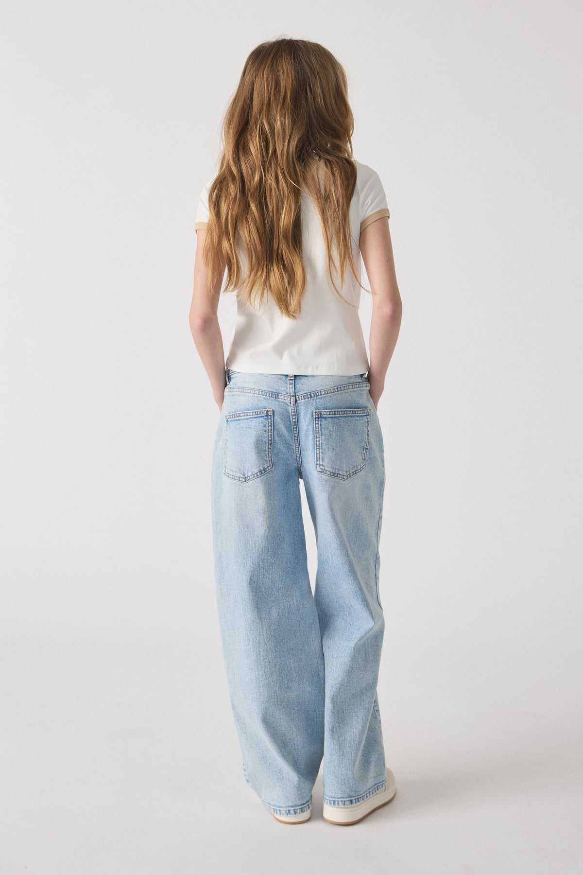 Girl facing away wearing a white ringer tee with beige trim and loose light wash denim jeans and white shoes.