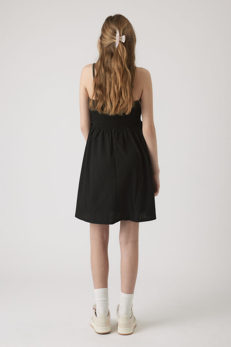 Girl wearing a black shirred dress with thin straps, styled with white socks, sneakers, and a hair clip in loose waves.