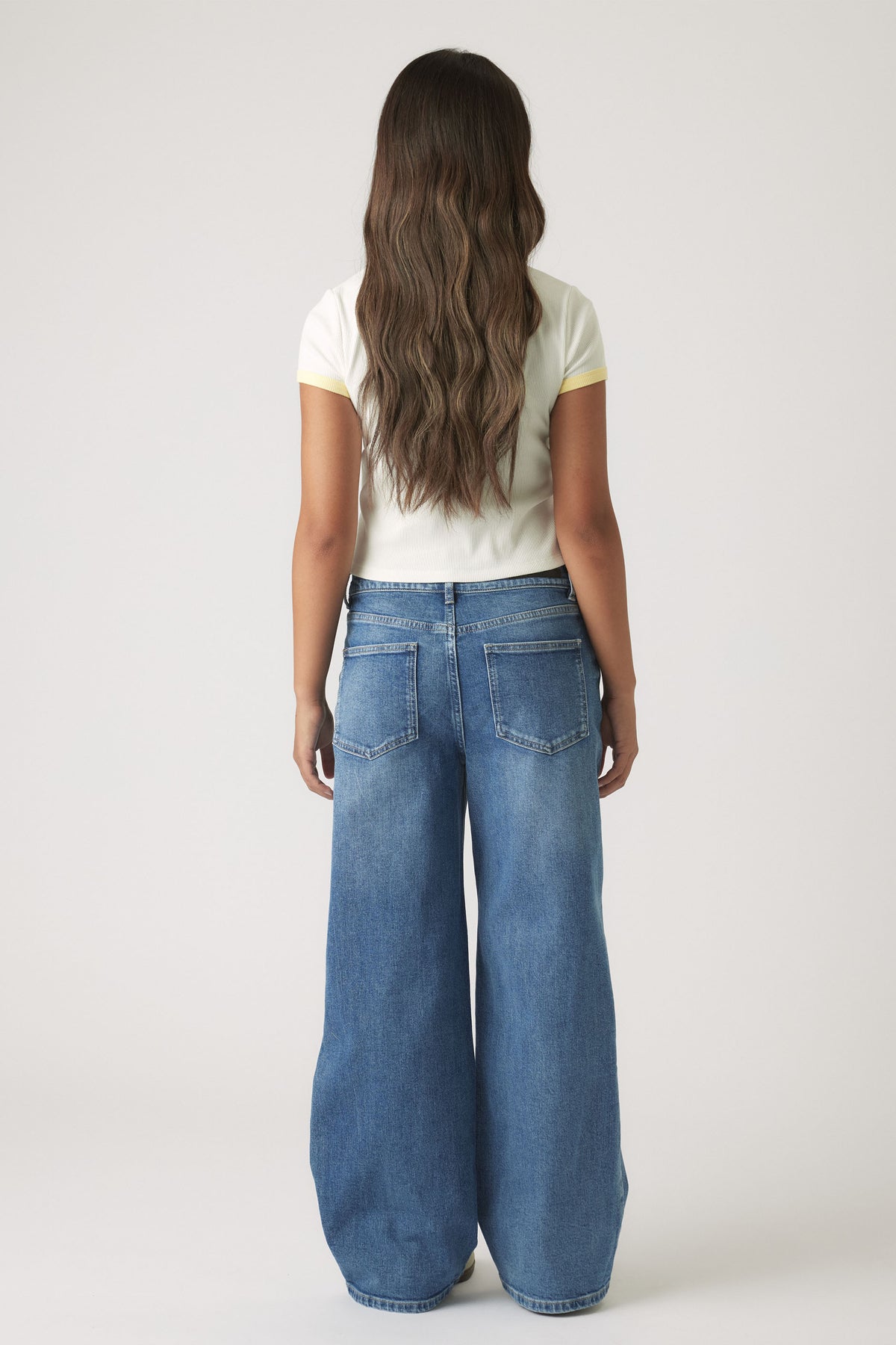 Back view of girl wearing a white cropped tee with yellow trim paired with wide-leg blue denim jeans