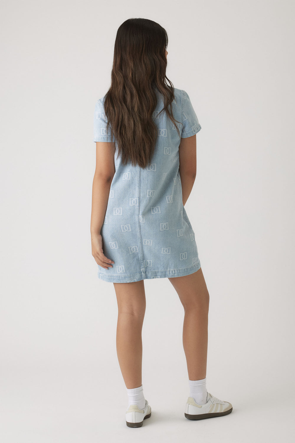 Girl wearing a light wash denim dress with short sleeves and subtle monogram print, paired with white socks and sneakers
