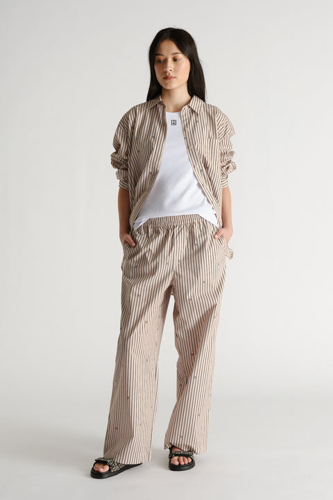Woman wearing beige monogram stripe pants and matching shirt over a white tee, styled with black sandals.
