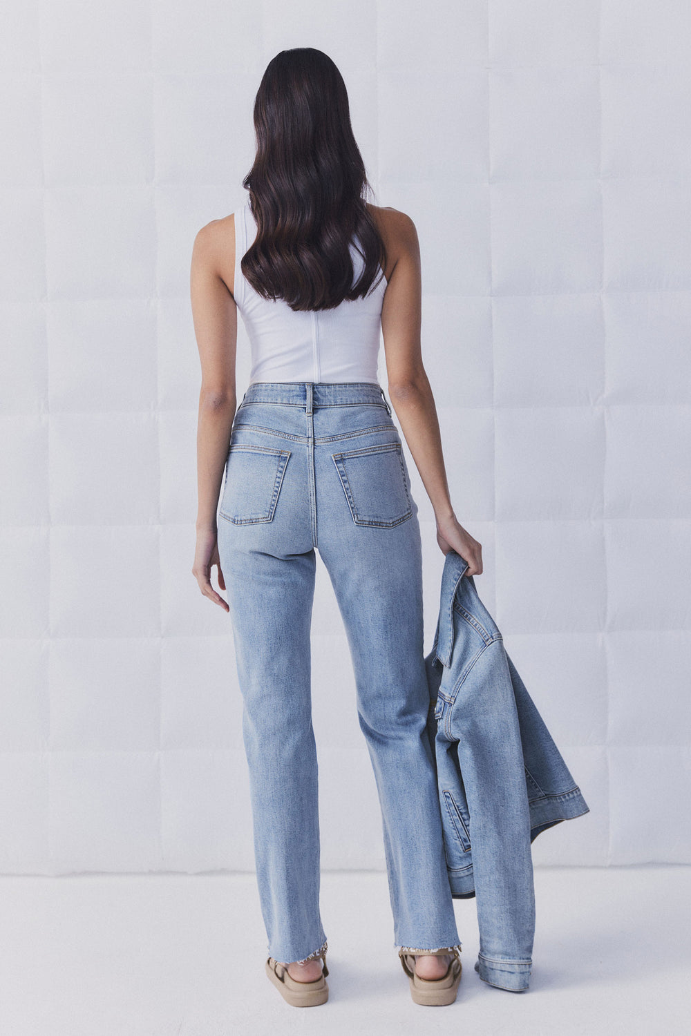 Woman seen from behind wearing light blue straight leg jeans and a white tank top, holding a matching denim jacket.
