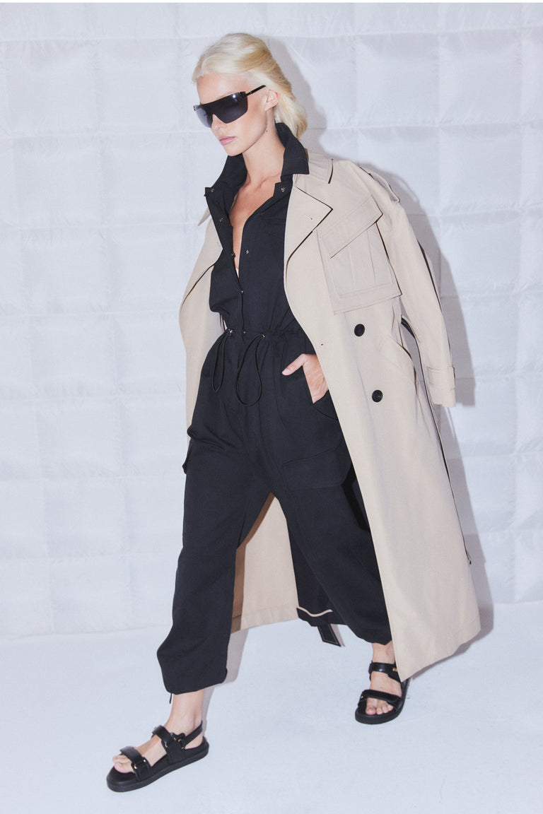 Woman in a black collared cargo jumpsuit with short sleeves and elastic waist, layered under a beige trench coat