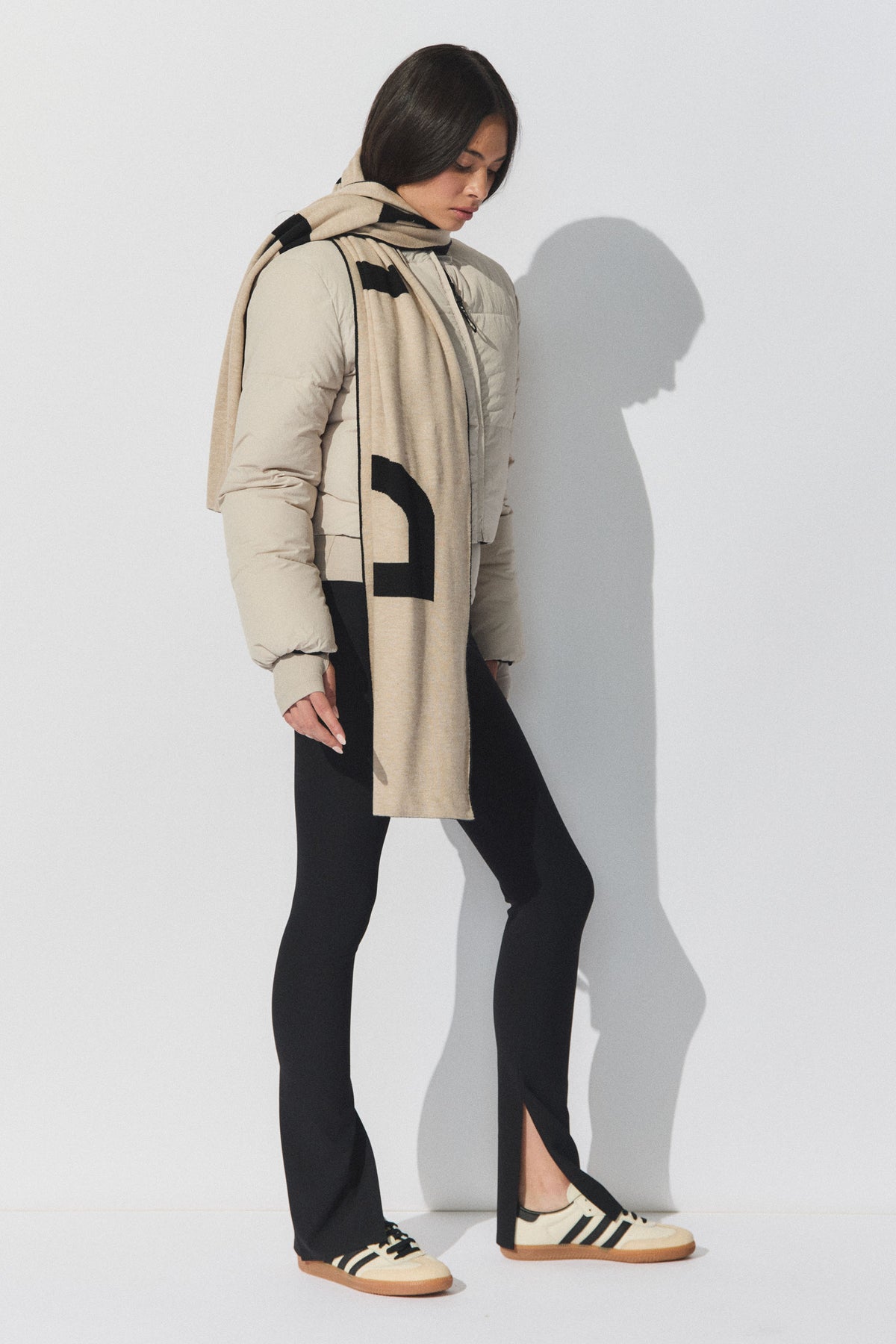 Woman wearing black ribbed leggings with front splits, styled with a beige puffer jacket, graphic scarf, and sneakers.