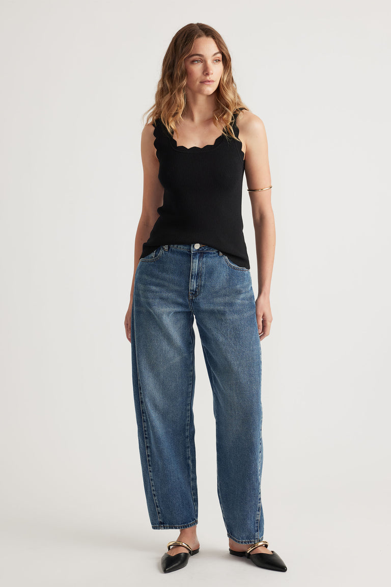 Woman wearing a black scallop edge tank top paired with relaxed fit blue denim jeans and black pointed flats.