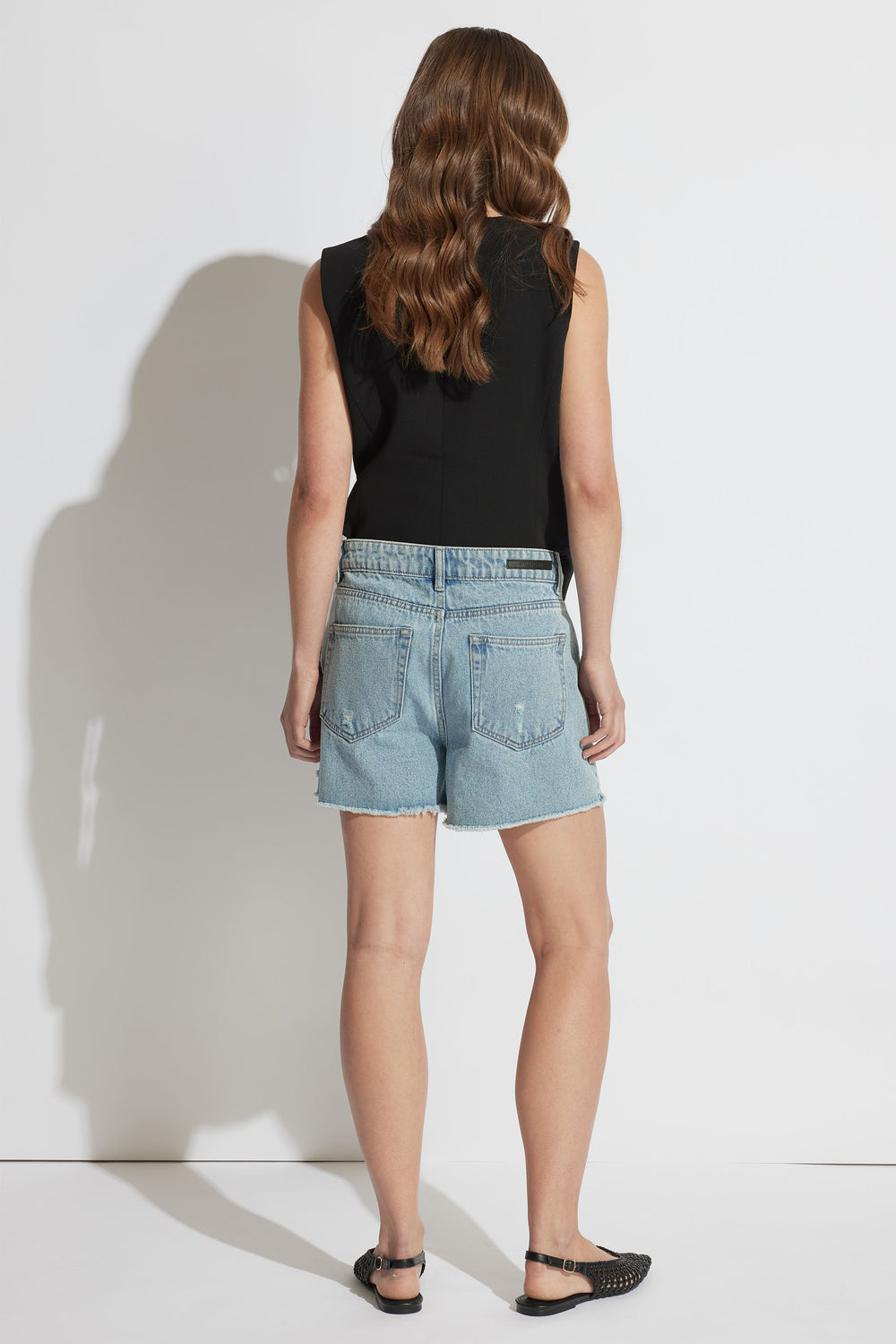 Back view of woman wearing light blue distressed denim shorts paired with a sleeveless black top and black sandals