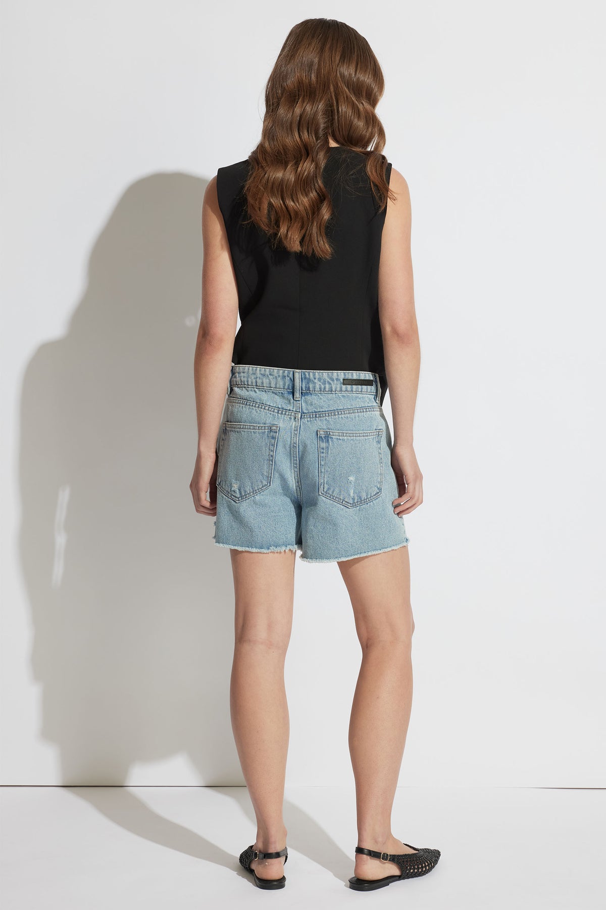 Back view of woman wearing light blue distressed denim shorts paired with a sleeveless black top and black sandals