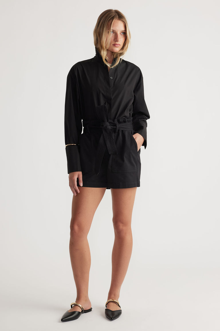 Woman wearing a belted black long-sleeve utility-style romper paired with pointed black flats featuring gold detail