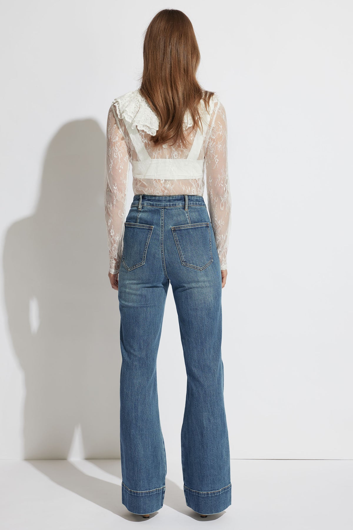 Woman wearing an ivory sheer lace top with ruffle detail paired with high-rise blue wide-leg jeans, back view.