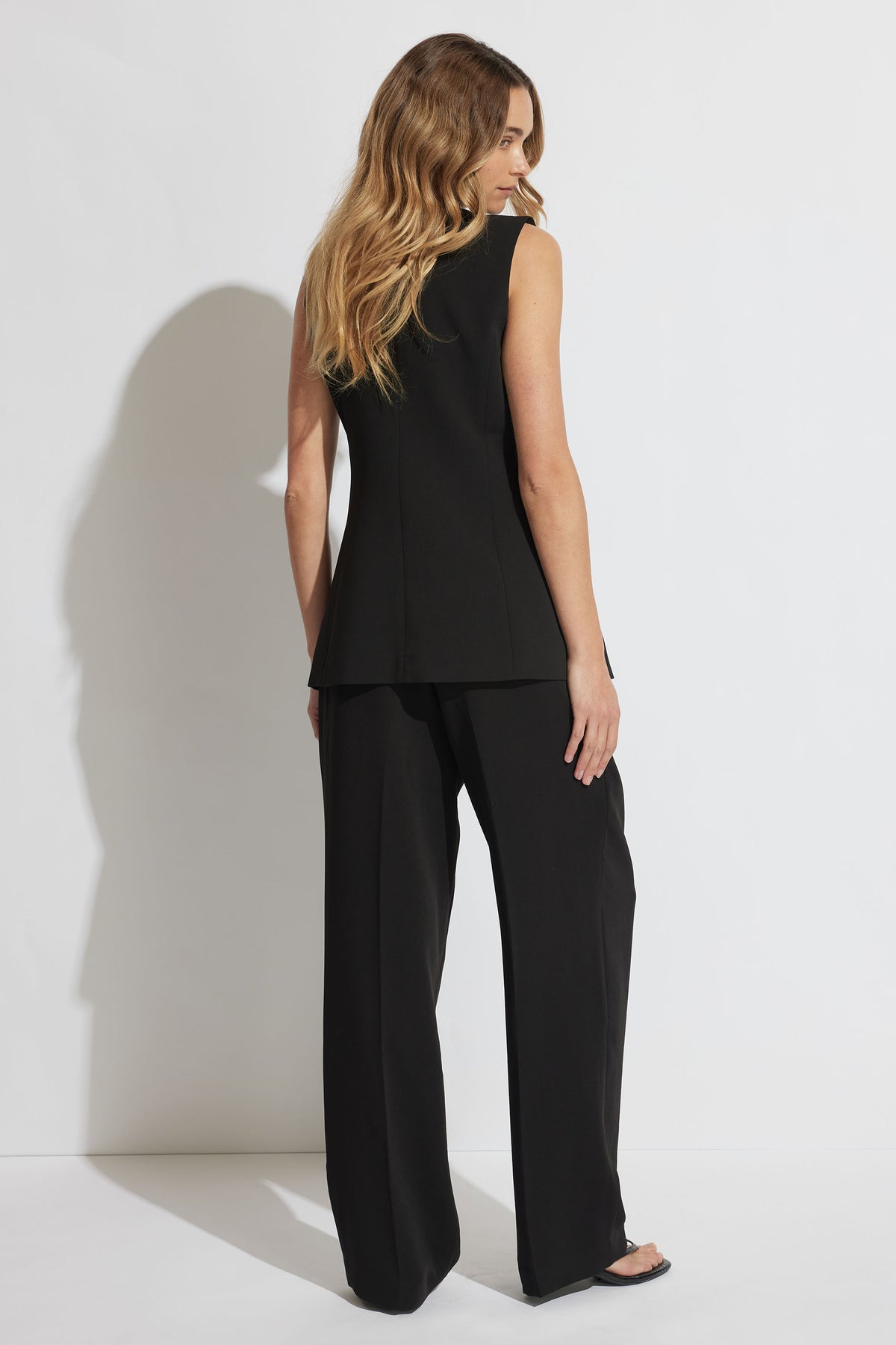 Woman wearing a tailored black sleeveless vest and wide-leg pants, shown from the back with loose waves in hair.