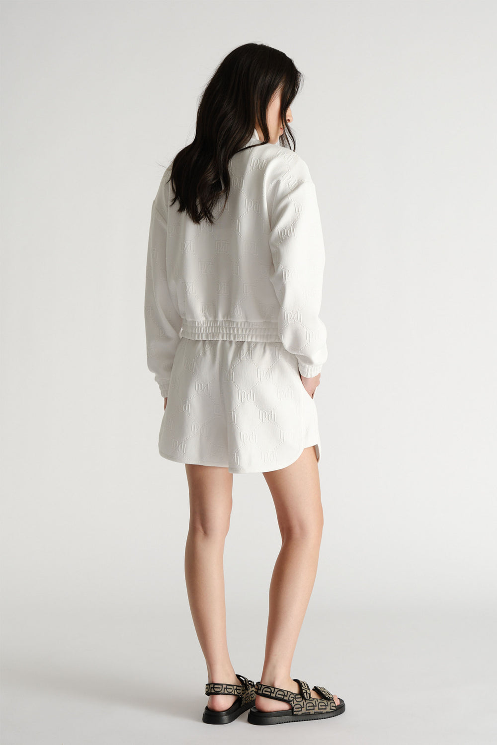 Back view of a woman wearing white embossed sweat shorts and matching jacket paired with black patterned sandals