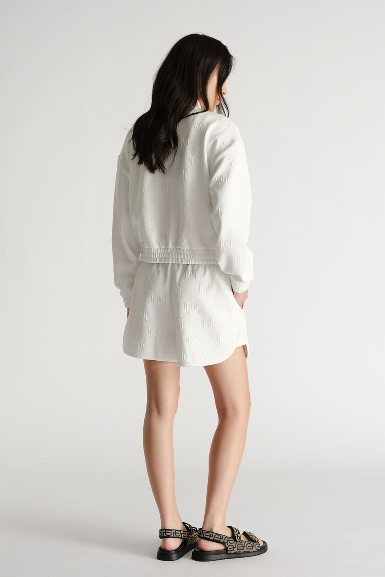 Back view of a woman wearing white embossed sweat shorts and matching jacket paired with black patterned sandals