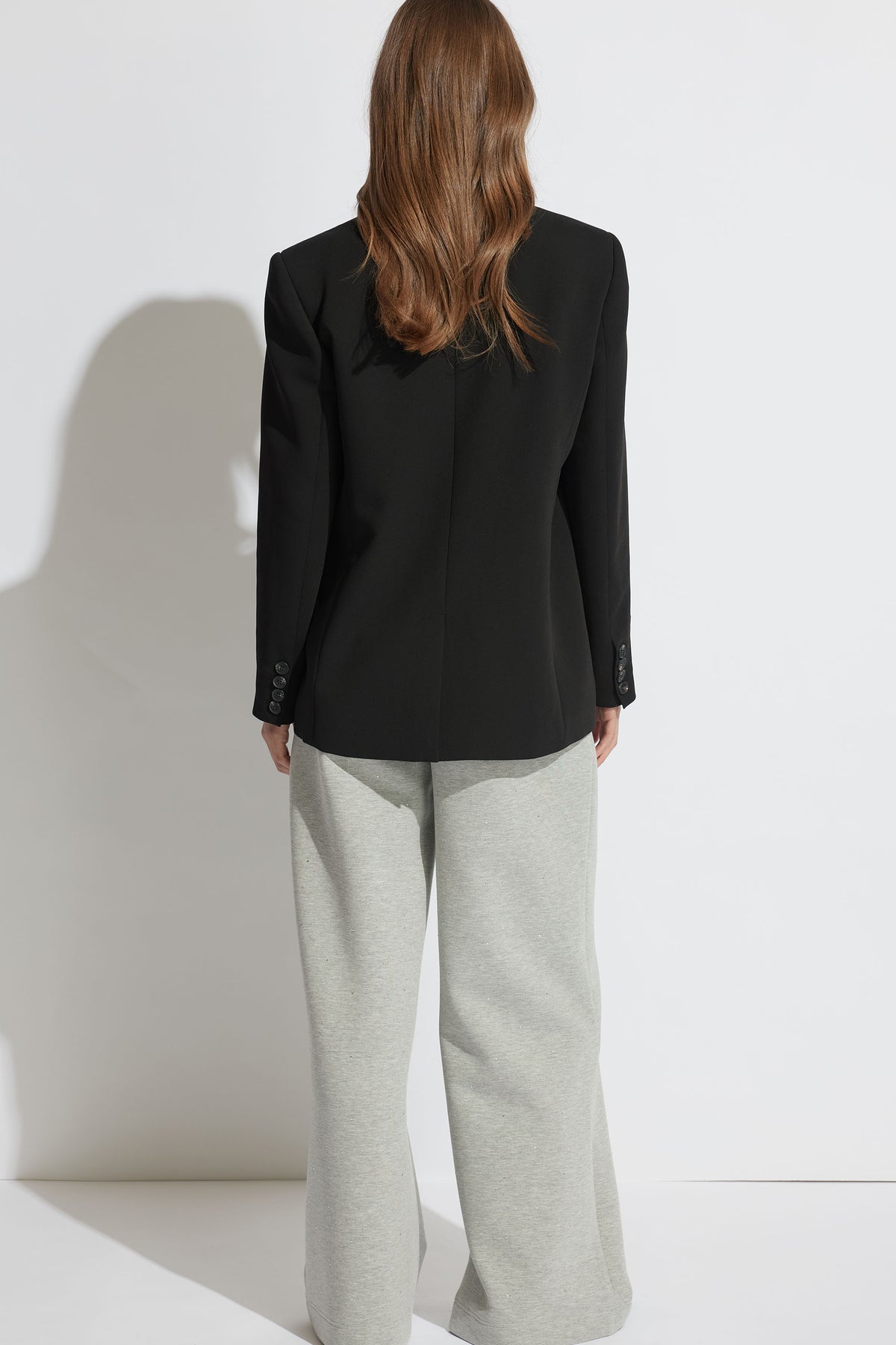 Woman with long hair wearing a black tailored blazer and light grey wide-leg pants, standing against a white wall.
