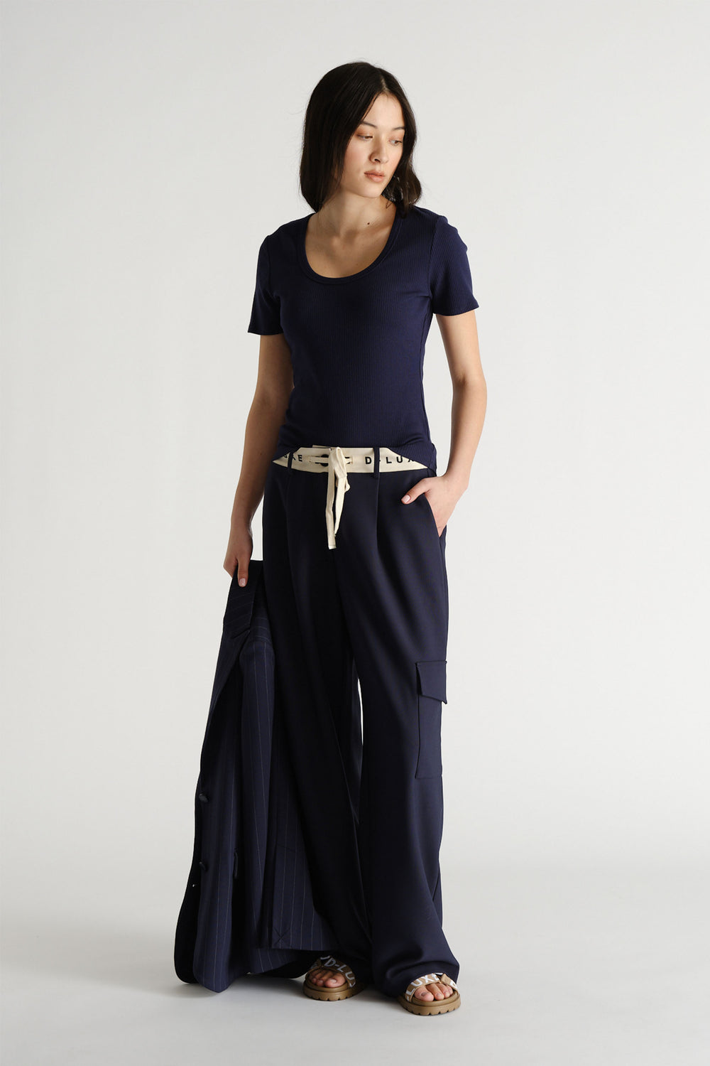 Woman wearing navy wide-leg pants with a white logo waistband, a fitted navy tee, and holding a matching jacket.